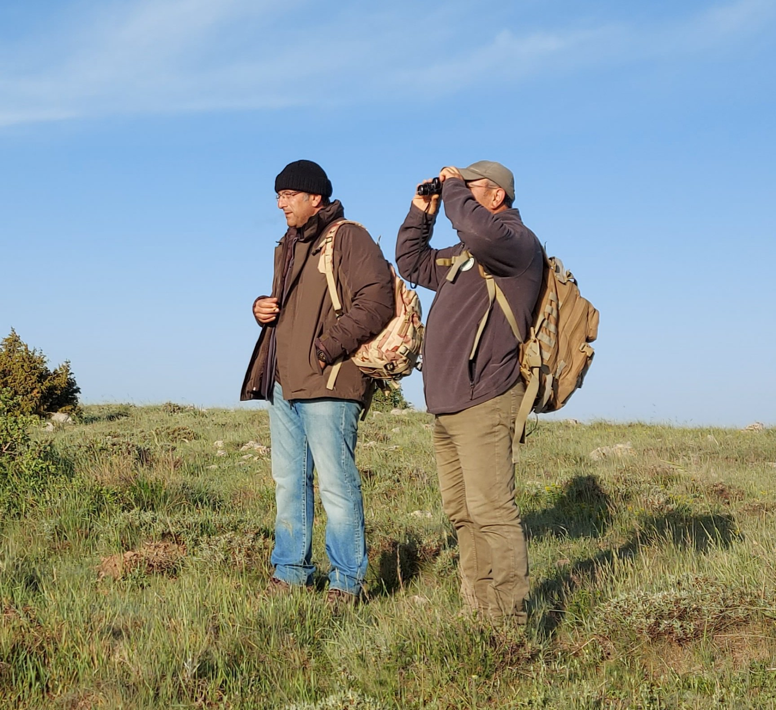 Two men standing in a field looking through binoculars
Description automatically generated